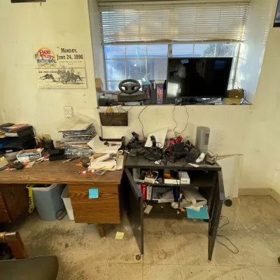 Sale Photo Thumbnail #56: Cluttered office area featuring a wooden desk, a dark grey cabinet with doors, electronics including a computer monitor, flight simulator joystick, and gaming steering wheel, alongside a framed 1996 Bob Feist Invitational poster.