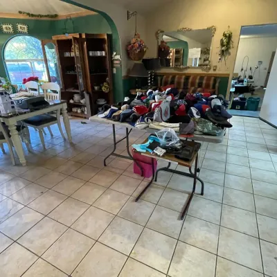 Sale Photo Thumbnail #94: A large collection of baseball caps and small accessories displayed on folding tables in a tiled room, with wooden hutches and a dining set in the background.