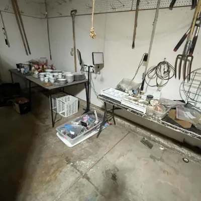 Sale Photo Thumbnail #79: Estate sale setup in a garage or basement featuring folding tables with dishware sets, craft supplies, and various yard tools on pegboard walls.