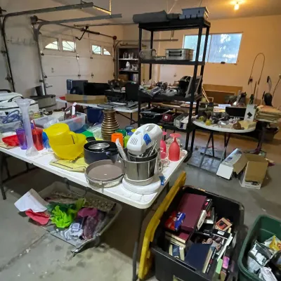 Sale Photo Thumbnail #69: Garage sale setup featuring folding tables and shelving units loaded with housewares, cookware, small appliances, and storage bins filled with books and media.