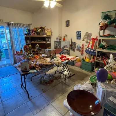 Sale Photo Thumbnail #45: A room filled with assorted estate sale merchandise, including folding tables laden with holiday decor, boxed items, home accessories, and shelving units with plush toys and craft supplies.
