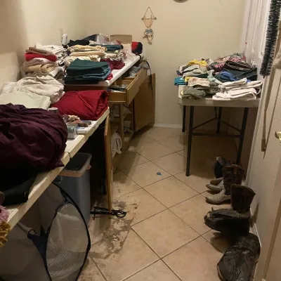 Sale Photo Thumbnail #84: A utility room filled with assorted linens, fabrics, and towels stacked on counters and folding tables, alongside several pairs of western cowboy boots lined up on the tiled floor.
