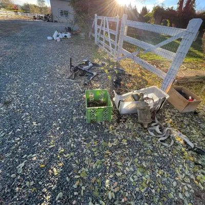 Sale Photo Thumbnail #30: Assorted yard and outdoor items, including a green plastic crate, clear plastic bin with vintage carriage-style lanterns, straps, and trailer hitch components on a gravel driveway.