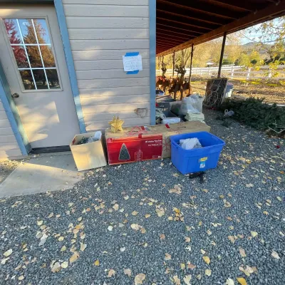 Sale Photo Thumbnail #39: Collection of outdoor Christmas decorations, including a boxed 9-foot Wesley Pine tree, a gold tree topper star, blue storage tote, and various string lights arranged along a gravel driveway.