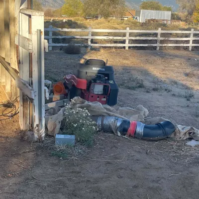Sale Photo Thumbnail #33: Red Snapper rear-engine riding lawn mower parked outdoors next to a shed, with attached vacuum chute hose, showing typical signs of outdoor storage and wear.
