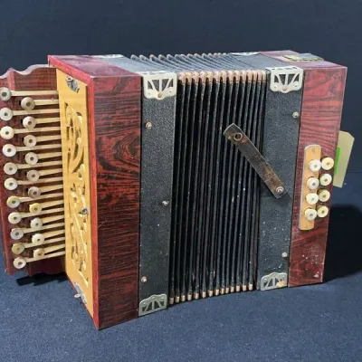 Sale Photo Thumbnail #34: Antique wooden button accordion featuring a faux rosewood finish, carved wood trim, metal corner brackets, and mother-of-pearl buttons.