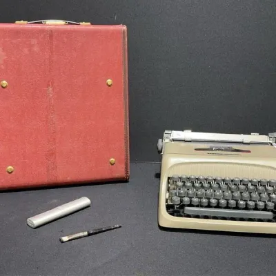 Sale Photo Thumbnail #37: Vintage Smith Corona manual typewriter in light beige with dark grey keys, displayed alongside a red carrying case, brush, and small accessory cylinder.