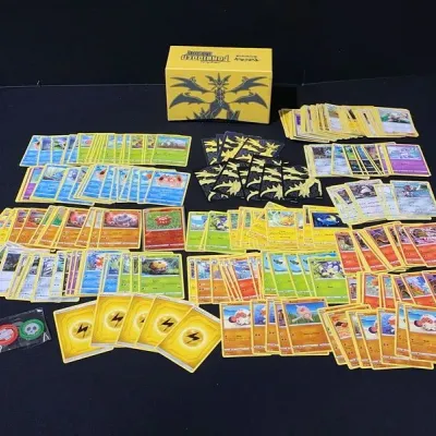 Sale Photo Thumbnail #74: Large collection of Pokémon trading cards sorted into piles, featuring a yellow Forbidden Light deck box, card sleeves, and gameplay tokens.