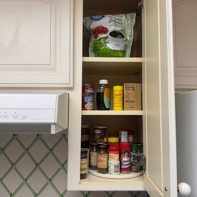 Sale Photo Thumbnail #23: Open kitchen cabinet stocked with pantry items, including stevia sweetener, canned goods, honey, and a lazy Susan filled with various spice jars.