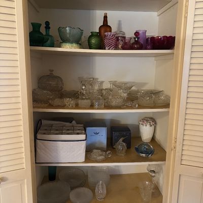 Sale Photo Thumbnail #44: A collection of vintage glassware, including colored glass vases, cut crystal bowls, and stemware arranged inside a white louvered-door cabinet.
