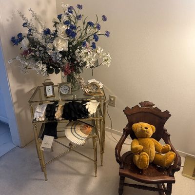 Sale Photo Thumbnail #53: A brass and glass nesting table display featuring small tabletop clocks, dress gloves, and a decorative fan beneath a large floral arrangement, alongside a small wooden child's rocking chair holding a plush teddy bear.