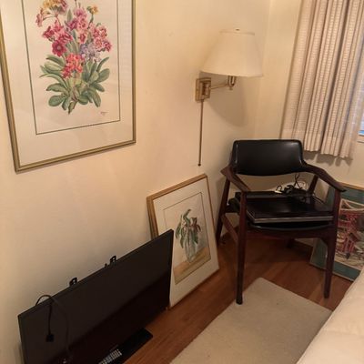 Sale Photo Thumbnail #61: A room corner featuring a mid-century modern armchair with black vinyl upholstery, framed floral artwork, a swing-arm wall sconce lamp, and a flat-screen TV.