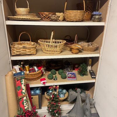 Sale Photo Thumbnail #78: A wooden cabinet filled with a variety of woven baskets on the top shelves and holiday decorations, including Christmas ornaments, miniature trees, and metal angel figures, on the lower shelves.