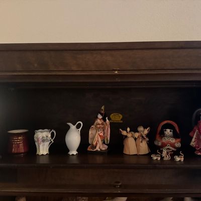 Sale Photo Thumbnail #45: A shelf inside a wooden cabinet featuring ceramic vases, Japanese geisha figurines, corn husk dolls, and miniature animal trinkets.