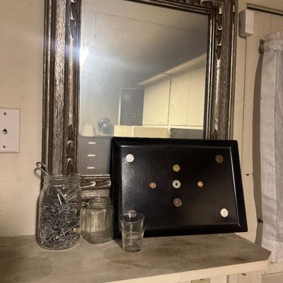 Sale Photo Thumbnail #75: Large mirror with a distressed silver-finished carved frame, displayed with a black decorative coin-embellished tray and glass jars filled with hardware.