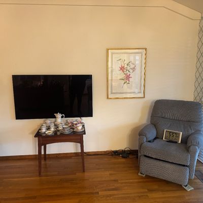 Sale Photo Thumbnail #39: Living room setup featuring a gray upholstered recliner, framed botanical wall art, a flat-screen television mounted above a small side table holding a teacup set, and polished hardwood floors.