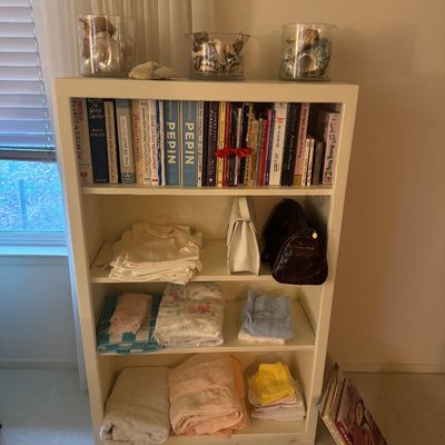 Sale Photo Thumbnail #54: White wooden four-shelf bookcase display featuring an assortment of hardbound books, seashells in glass vases, handbags, and folded linens.