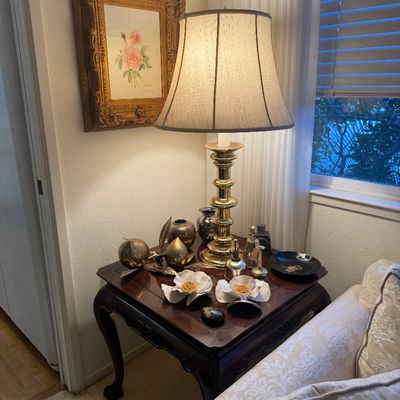 Sale Photo Thumbnail #17: Traditional brass table lamp with a fabric bell shade, sitting on an Asian-style dark wood end table alongside decorative brass fruit, ceramic flowers, and small vases.