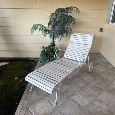 Sale Photo Thumbnail #93: Vintage white wrought iron slatted lounge chair with scrollwork armrests and rear wheels, shown alongside a potted faux palm plant.