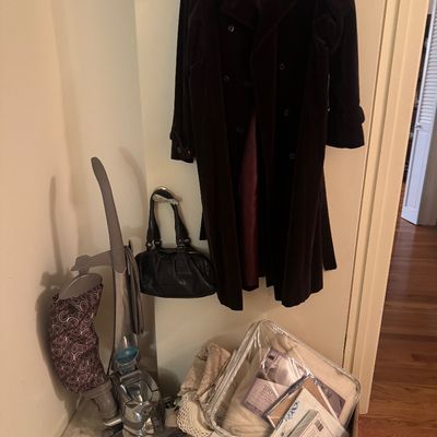 Sale Photo Thumbnail #58: Dark purple double-breasted long coat hanging beside a black leather handbag, upright vacuum, and a box of packaged linens.