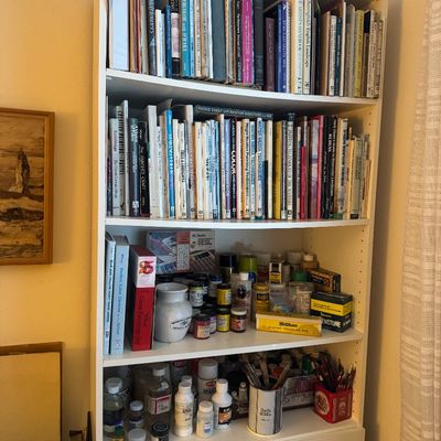 Sale Photo Thumbnail #68: Tall white wooden bookcase filled with art instruction books, paints, brushes, and various art supplies.