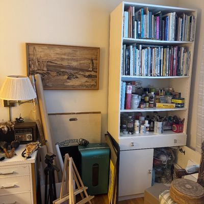 Sale Photo Thumbnail #63: Tall white painted bookshelf filled with books and art supplies, alongside a wooden easel, green paper shredder, framed landscape artwork, and small white side table.