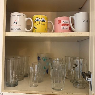 Sale Photo Thumbnail #25: Kitchen cabinet filled with ceramic novelty mugs, including a SpongeBob SquarePants mug, alongside an assortment of clear drinking glasses and glass beer steins.