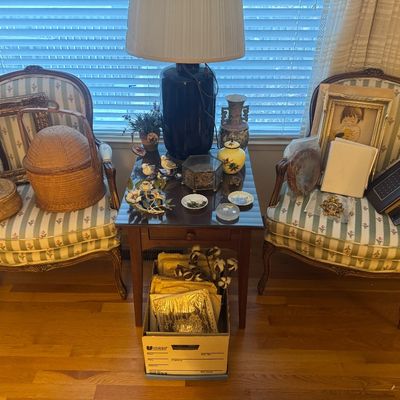 Sale Photo Thumbnail #67: Pair of French provincial-style armchairs with floral stripe upholstery, flanking a wooden side table displaying a large dark lamp, small Asian ceramics, woven baskets, framed artwork, and decorative trinkets.
