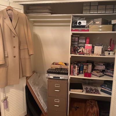 Sale Photo Thumbnail #60: Closet filled with diverse household items, including a camel hair overcoat hanging on the door, a two-drawer metal filing cabinet, electronics, books, media, and storage boxes.