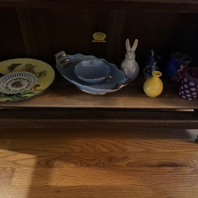 Sale Photo Thumbnail #48: Collection of vintage decorative glassware and porcelain items, including plates, a light blue dish set, a white rabbit figurine, and colorful small vases displayed inside a wooden cabinet shelf.