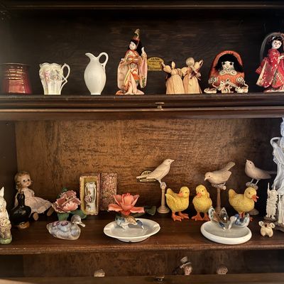 Sale Photo Thumbnail #50: Collection of vintage figurines displayed inside a wooden bookcase, including Japanese geisha dolls, porcelain birds, ceramic ducks, miniature vases, and small decorative sculptures.