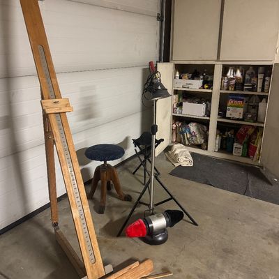 Sale Photo Thumbnail #74: Tall wooden artist floor easel accompanied by a blue cushioned swivel stool, adjustable clip light on a tripod stand, and additional art supplies.
