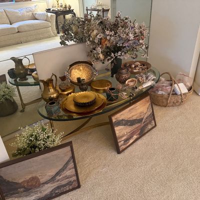 Sale Photo Thumbnail #11: Glass-top oval console table displaying brass pitchers, decorative plates, sea shells, dried floral arrangements, and framed landscape artwork.