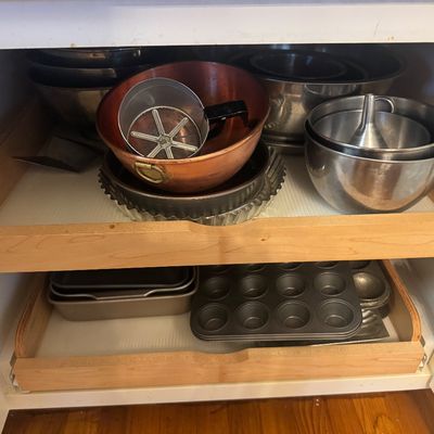 Sale Photo Thumbnail #36: Assortment of metal bakeware and kitchen supplies stored in pull-out cabinet shelves, including stainless steel mixing bowls, a copper basin, flour sifter, pie pans, and muffin tins.