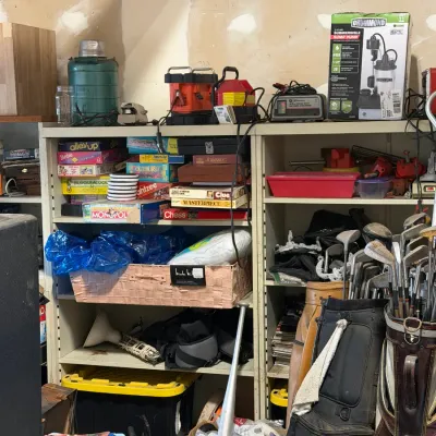 Sale Photo Thumbnail #4: Metal shelving unit packed with vintage board games, battery chargers, a boxed Drummond sump pump, golf bags with clubs, and various household storage items.