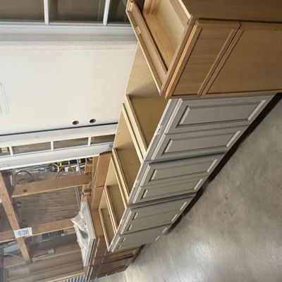 Sale Photo Thumbnail #80: Row of unfinished and painted wood kitchen base and wall cabinets lined up on a warehouse floor alongside a white exterior door.