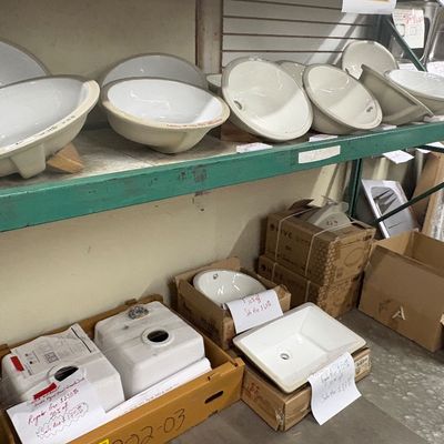 Sale Photo Thumbnail #85: Collection of white porcelain undermount and drop-in bathroom sinks displayed on green warehouse shelving, both loose and in original cardboard boxes with handwritten price tags.
