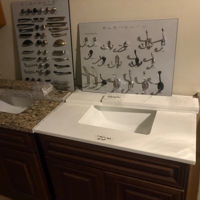 Sale Photo Thumbnail #13: Bathroom vanities with stone countertops and undermount sinks, displayed alongside sample boards of Elements brand cabinet hardware and hooks.