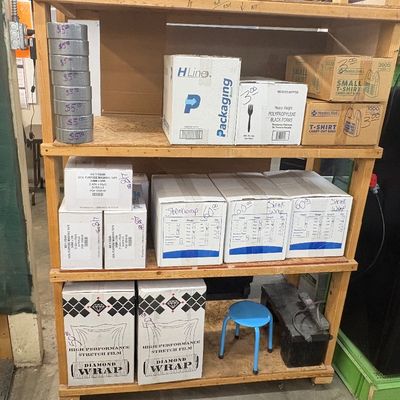 Sale Photo Thumbnail #71: Wooden utility shelving unit stocked with various packaging supplies, including duct tape, shrink wrap, stretch film, masking tape, and Member's Mark t-shirt carry-out bags.