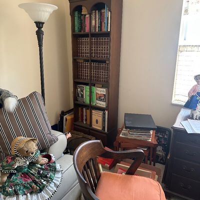 Sale Photo Thumbnail #71: Tall dark wood bookcase filled with hardback book sets, situated beside a floor lamp, striped armchair, and a vintage wooden banker's chair with an orange cushion.