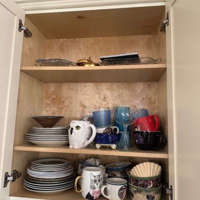 Sale Photo Thumbnail #40: Open kitchen cabinet filled with diverse dishware, novelty mugs including an owl and cat illustration, glassware, plates, and serving bowls.