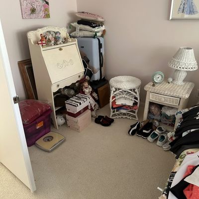 Sale Photo Thumbnail #51: A bedroom area featuring a vintage white drop-front secretary desk, white wicker nightstand and accent stool, a white wicker lamp, framed wall art, and various personal decor items.