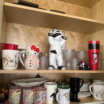 Sale Photo Thumbnail #38: A collection of novelty mugs and glassware displayed inside a wooden cabinet, featuring a mummy cat figurine, a Hello Kitty snowman mug, and Betty Boop cups.