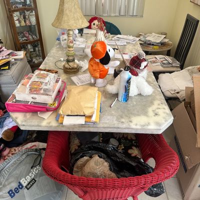 Sale Photo Thumbnail #86: Faux marble dining table cluttered with various household goods, featuring a painted ceramic table lamp, plush toys, and a bright red wicker tub chair.