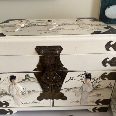 Sale Photo Thumbnail #78: Vintage Asian cream lacquer jewelry chest featuring mother-of-pearl or soapstone geisha figure relief inlays, brass corner brackets, and a decorative front latch.