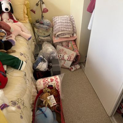 Sale Photo Thumbnail #81: Collection of plush toys, soft throw blankets, and packaged linens stacked beside a bed, including a Snoopy plush and a small floral lamp.