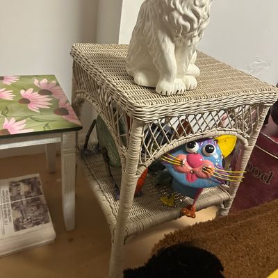 Sale Photo Thumbnail #20: White wicker two-tiered side table featuring a large ceramic cat statue on top and a colorful whimsical metal cat figurine on the lower shelf.