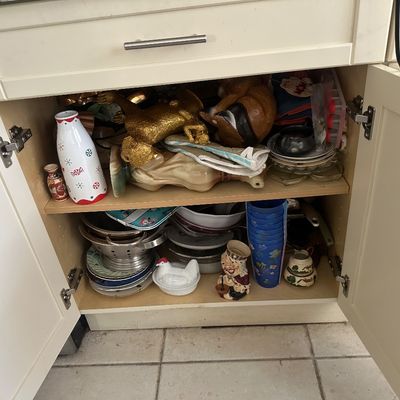 Sale Photo Thumbnail #39: Open kitchen cabinet filled with assorted housewares, including decorative ceramics, stacked blue plastic cups, cookware, a colander, and a white milk glass hen on a nest dish.