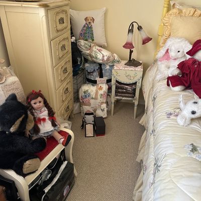 Sale Photo Thumbnail #79: Bedroom corner featuring an off-white French Provincial-style tall dresser, a floral painted nightstand with a lily lamp, folded throw blankets, and various stuffed animals and dolls.