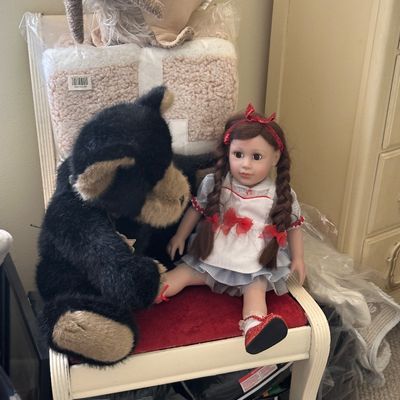Sale Photo Thumbnail #84: Collection of plush toys and a porcelain Dorothy-style doll seated on a white upholstered vanity stool, with packaged blankets stacked behind them.